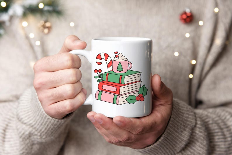 May include: A white ceramic mug featuring a colourful graphic of a stack of three books in green, red, and pink. A small pink mug with a green tree icon, a candy cane, and holly berries with green leaves are illustrated on top of the books.