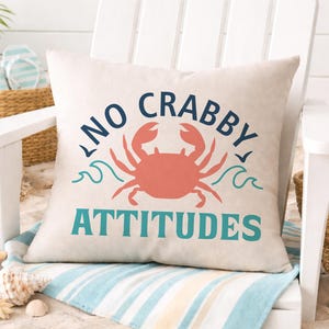 Puede incluir: Un coj&iacute;n cuadrado beige con las palabras "NO CRABBY ATTITUDES" en azul marino y verde azulado, y una ilustraci&oacute;n de cangrejo coral. El coj&iacute;n est&aacute; sobre una silla blanca, con decoraci&oacute;n de tem&aacute;tica playera en el fondo.