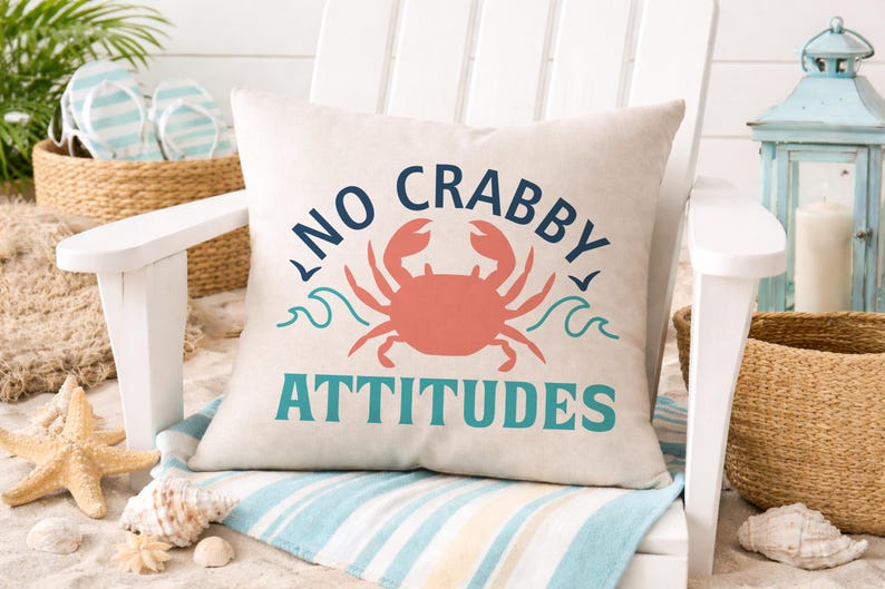 Puede incluir: Un coj&iacute;n cuadrado beige con las palabras "NO CRABBY ATTITUDES" en azul marino y verde azulado, y una ilustraci&oacute;n de cangrejo coral. El coj&iacute;n est&aacute; sobre una silla blanca, con decoraci&oacute;n de tem&aacute;tica playera en el fondo.