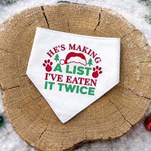 Op de afbeelding: Witte hondenbandana met de tekst "He's making a list, I've eaten it twice" in rood en groen. Het ontwerp bevat een kerstmuts, pootafdrukken en kerstbomen. Op een houten ondergrond met kerstversieringen.