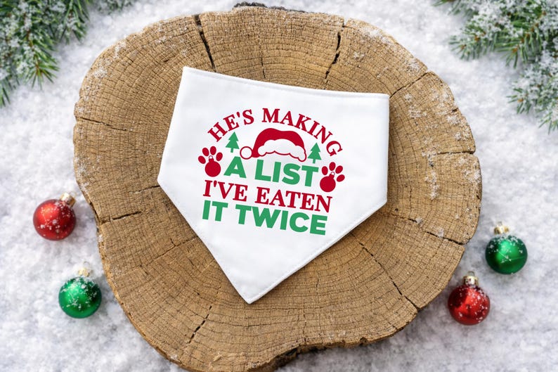 Op de afbeelding: Witte hondenbandana met de tekst "He's making a list, I've eaten it twice" in rood en groen. Het ontwerp bevat een kerstmuts, pootafdrukken en kerstbomen. Op een houten ondergrond met kerstversieringen.