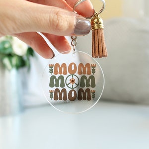 May include: A clear acrylic keychain with a brown tassel. The keychain features the word "MOM" repeated three times in a retro font with a peace sign in the center.