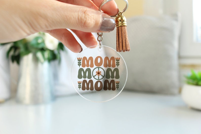 May include: A clear acrylic keychain with a brown tassel. The keychain features the word "MOM" repeated three times in a retro font with a peace sign in the center.