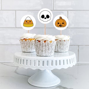 Pu&ograve; includere: Tre cupcake bianchi con glassa bianca sormontati da decorazioni per cupcake a tema Halloween. Le decorazioni sono un mais dolce, un teschio e una zucca. I cupcake sono su un supporto per torte bianco.