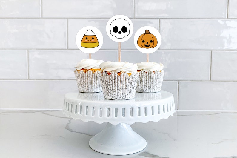 Pu&ograve; includere: Tre cupcake bianchi con glassa bianca sormontati da decorazioni per cupcake a tema Halloween. Le decorazioni sono un mais dolce, un teschio e una zucca. I cupcake sono su un supporto per torte bianco.