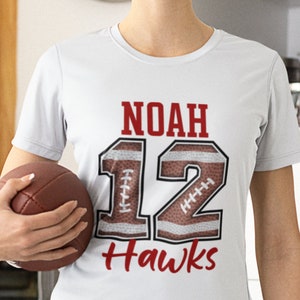 Puede incluir: Camiseta blanca con el nombre "NOAH", el n&uacute;mero "12" y la palabra "Hawks" en rojo. El n&uacute;mero "12" est&aacute; dise&ntilde;ado para parecerse a un bal&oacute;n de f&uacute;tbol americano. Un bal&oacute;n de f&uacute;tbol americano marr&oacute;n se sostiene en la mano.