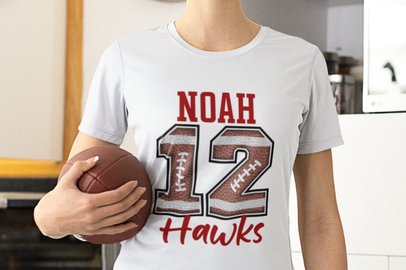 Puede incluir: Camiseta blanca con el nombre "NOAH", el n&uacute;mero "12" y la palabra "Hawks" en rojo. El n&uacute;mero "12" est&aacute; dise&ntilde;ado para parecerse a un bal&oacute;n de f&uacute;tbol americano. Un bal&oacute;n de f&uacute;tbol americano marr&oacute;n se sostiene en la mano.