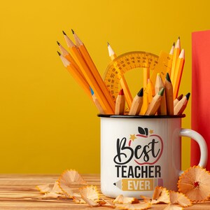 May include: A white enamel mug with the text "Best Teacher Ever" filled with pencils, a protractor, and a ruler. Two books, one red and one yellow, stand behind the mug. Pencil shavings are scattered on the wooden surface.