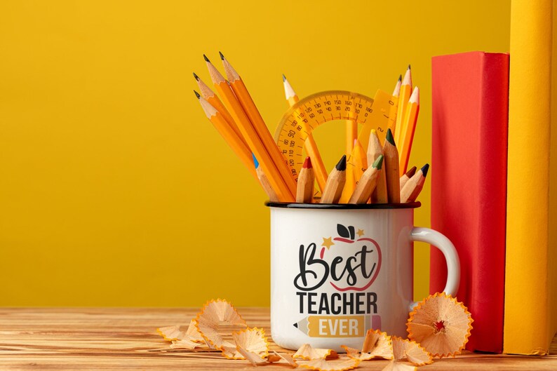 May include: A white enamel mug with the text "Best Teacher Ever" filled with pencils, a protractor, and a ruler. Two books, one red and one yellow, stand behind the mug. Pencil shavings are scattered on the wooden surface.