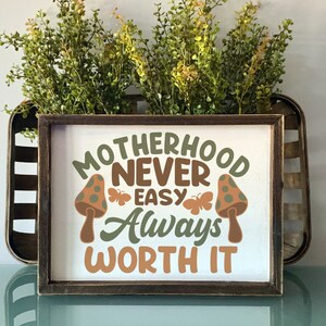 May include: A wooden sign with the text "Motherhood Never Easy Always Worth It" in brown, green, and orange lettering. The sign is decorated with two mushroom illustrations with green and brown polka dots.