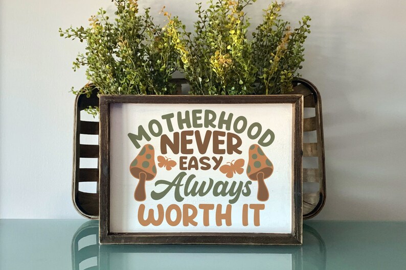 May include: A wooden sign with the text "Motherhood Never Easy Always Worth It" in brown, green, and orange lettering. The sign is decorated with two mushroom illustrations with green and brown polka dots.