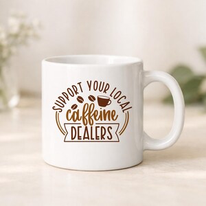 May include: White ceramic coffee mug with a brown graphic that reads "Support Your Local Caffeine Dealers." The design includes coffee beans and a coffee cup. The mug has a curved handle and is set against a neutral background.