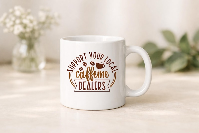 May include: White ceramic coffee mug with a brown graphic that reads "Support Your Local Caffeine Dealers." The design includes coffee beans and a coffee cup. The mug has a curved handle and is set against a neutral background.