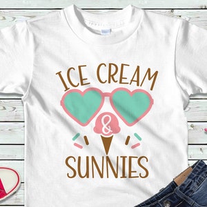 May include: White t-shirt with the text "ICE CREAM & SUNNIES" in brown letters. The text is surrounded by a pink and green heart-shaped sunglasses graphic and a pink and white ice cream cone graphic.