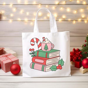 May include: A white tote bag featuring a graphic illustration of a stack of three books topped with a pink mug of hot cocoa and a candy cane. The design includes green and pink book covers, holly leaves, and red berries in a festive style.