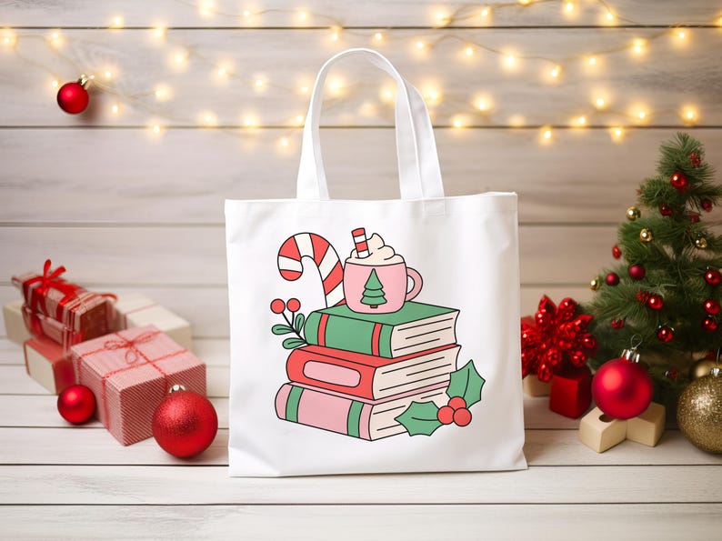 May include: A white tote bag featuring a graphic illustration of a stack of three books topped with a pink mug of hot cocoa and a candy cane. The design includes green and pink book covers, holly leaves, and red berries in a festive style.