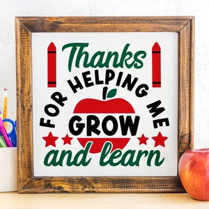 May include: A framed sign with the text "Thanks for helping me grow and learn" in green and black, with a red apple and crayons. A red apple sits to the right of the frame. A pencil holder with pens and pencils is to the left.