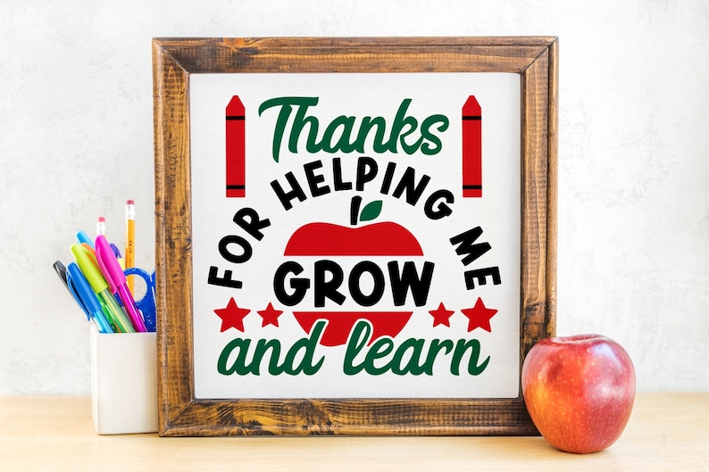 May include: A framed sign with the text "Thanks for helping me grow and learn" in green and black, with a red apple and crayons. A red apple sits to the right of the frame. A pencil holder with pens and pencils is to the left.