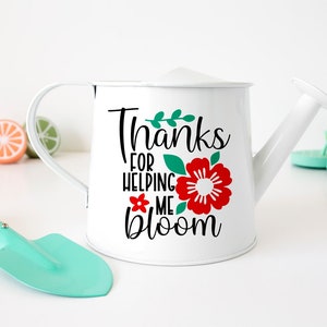 Puede incluir: Regadera de metal blanco con asa y boquilla curvadas. La regadera presenta el texto "Thanks for helping me bloom" con una flor roja y hojas verdes. Una peque&ntilde;a paleta turquesa y fruta de juguete en el fondo.