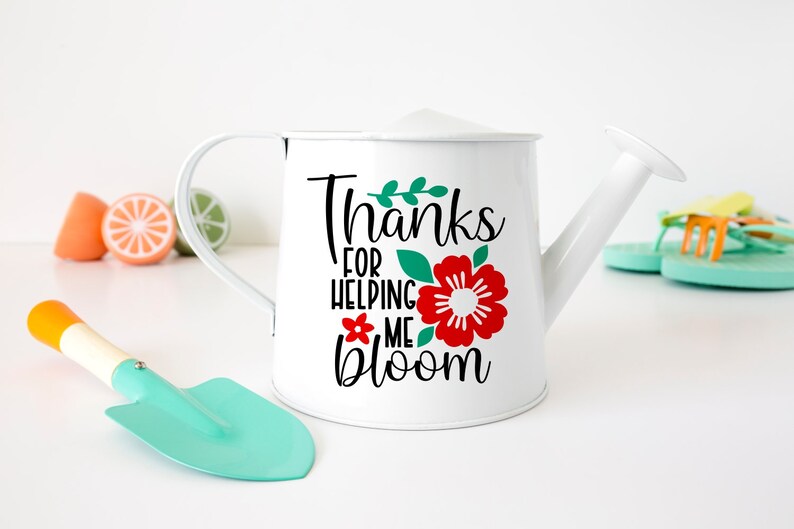 Puede incluir: Regadera de metal blanco con asa y boquilla curvadas. La regadera presenta el texto "Thanks for helping me bloom" con una flor roja y hojas verdes. Una peque&ntilde;a paleta turquesa y fruta de juguete en el fondo.