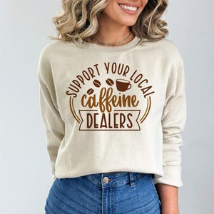 May include: A beige sweatshirt with the text "SUPPORT YOUR LOCAL caffeine DEALERS" in brown lettering. The design includes coffee beans and a coffee cup. The sweatshirt is made of a soft material.