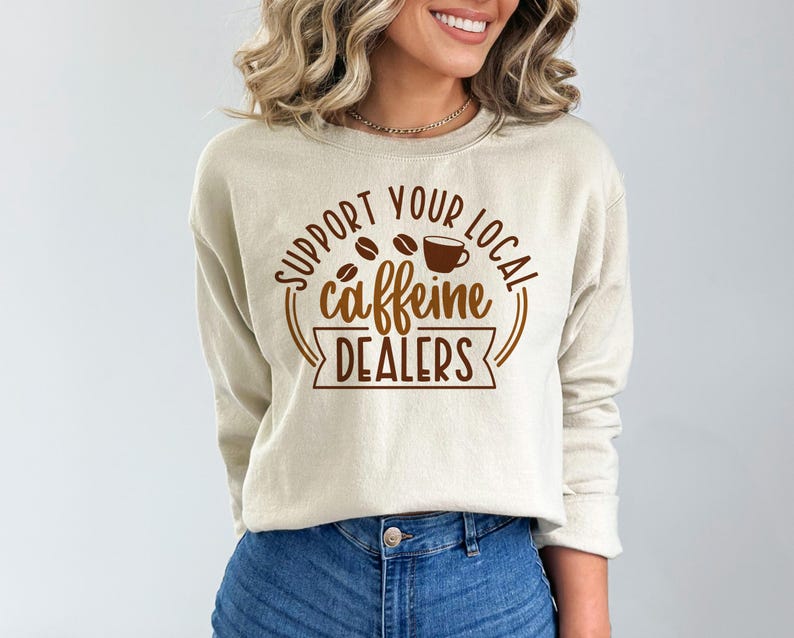 May include: A beige sweatshirt with the text "SUPPORT YOUR LOCAL caffeine DEALERS" in brown lettering. The design includes coffee beans and a coffee cup. The sweatshirt is made of a soft material.