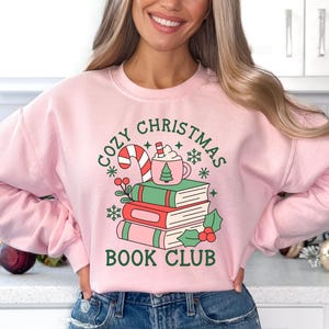 May include: A light pink crewneck sweatshirt featuring a graphic print of three stacked books, a candy cane, and a mug of hot cocoa. The text reads "COZY CHRISTMAS BOOK CLUB" in dark green lettering, surrounded by small snowflake and holly illustrations.