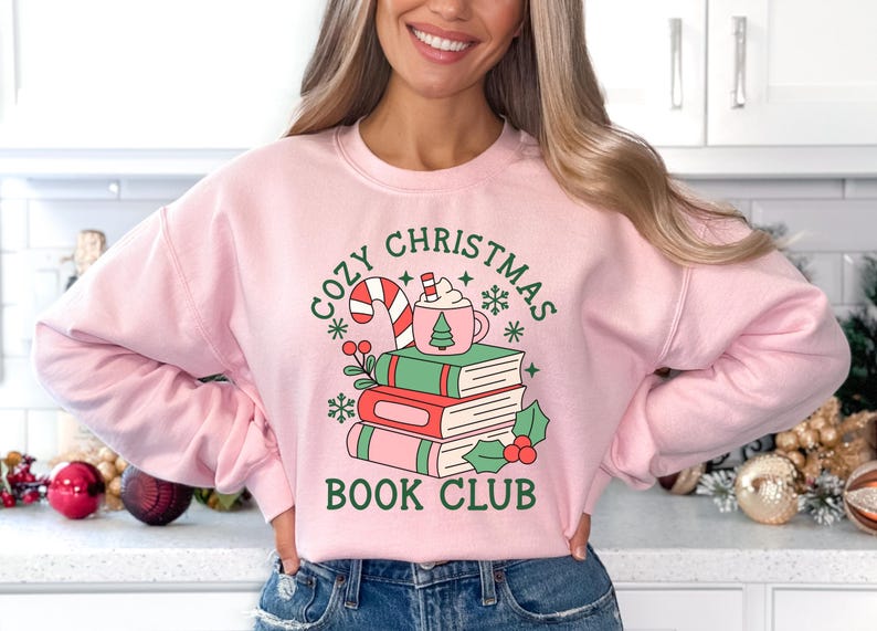 May include: A light pink crewneck sweatshirt featuring a graphic print of three stacked books, a candy cane, and a mug of hot cocoa. The text reads "COZY CHRISTMAS BOOK CLUB" in dark green lettering, surrounded by small snowflake and holly illustrations.