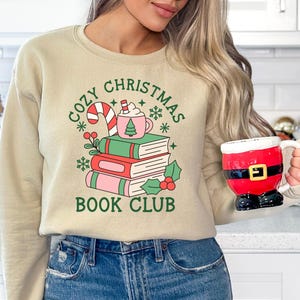 May include: A beige crewneck sweatshirt featuring a graphic of three stacked books, a candy cane, and a pink mug with a tree icon. The text reads COZY CHRISTMAS BOOK CLUB in green. A red and black Santa-themed mug is held in one hand.