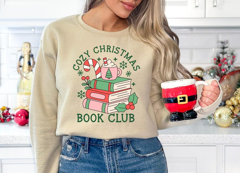 May include: A beige crewneck sweatshirt featuring a graphic of three stacked books, a candy cane, and a pink mug with a tree icon. The text reads COZY CHRISTMAS BOOK CLUB in green. A red and black Santa-themed mug is held in one hand.