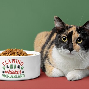 May include: White ceramic cat food bowl with the text "Clawing in a whisker wonderland" in red and green. A calico cat is sitting in front of the bowl on a red surface.