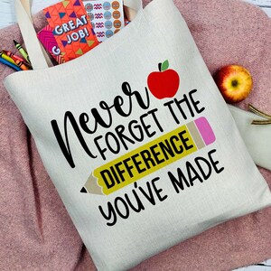 Puede incluir: Bolso tote color crema con el texto "Never Forget The Difference You've Made" y un gr&aacute;fico de manzana roja y l&aacute;piz. La bolsa est&aacute; llena de &uacute;tiles escolares y descansa sobre un su&eacute;ter rosa.