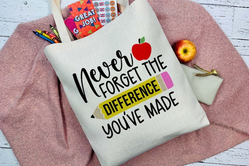 Puede incluir: Bolso tote color crema con el texto "Never Forget The Difference You've Made" y un gr&aacute;fico de manzana roja y l&aacute;piz. La bolsa est&aacute; llena de &uacute;tiles escolares y descansa sobre un su&eacute;ter rosa.