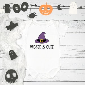 Pu&ograve; includere: Un body bianco per neonato con un cappello da strega viola e il testo "Wicked & Cute". Il body &egrave; circondato da decorazioni di Halloween, tra cui fantasmi, una zucca e un ragno.