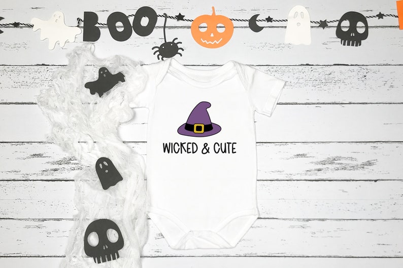 Pu&ograve; includere: Un body bianco per neonato con un cappello da strega viola e il testo "Wicked & Cute". Il body &egrave; circondato da decorazioni di Halloween, tra cui fantasmi, una zucca e un ragno.