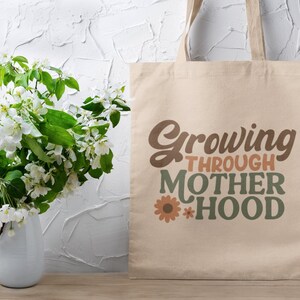 May include: A beige tote bag with the text "Growing Through Motherhood" in brown, orange, and green lettering with a small flower graphic.