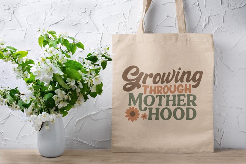 May include: A beige tote bag with the text "Growing Through Motherhood" in brown, orange, and green lettering with a small flower graphic.