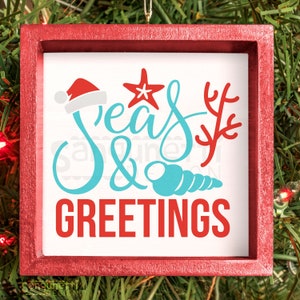 May include: A square, red-framed Christmas ornament with the words "Seas & Greetings" in blue and red script. The design includes a Santa hat, starfish, coral, and seashell. The ornament is hanging on a green, leafy background.