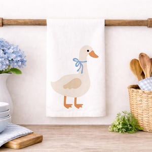 May include: A white kitchen towel with a cartoon goose wearing a blue bow. The towel hangs from a wooden rod. A white vase with blue flowers, a stack of bowls, and a basket with wooden spoons are also visible.