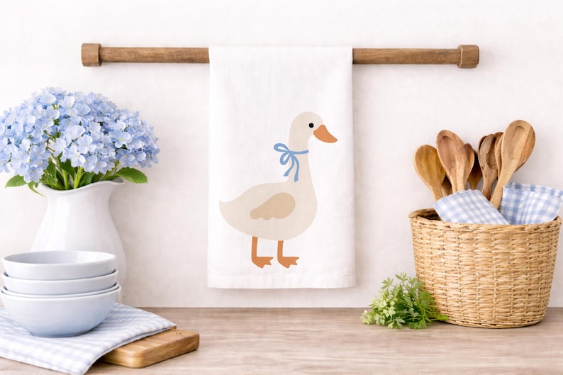 May include: A white kitchen towel with a cartoon goose wearing a blue bow. The towel hangs from a wooden rod. A white vase with blue flowers, a stack of bowls, and a basket with wooden spoons are also visible.