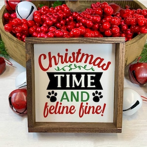 May include: A wooden sign with the text "Christmas Time And Feline Fine!" in red, green, and black lettering. The sign is surrounded by red and white jingle bells.