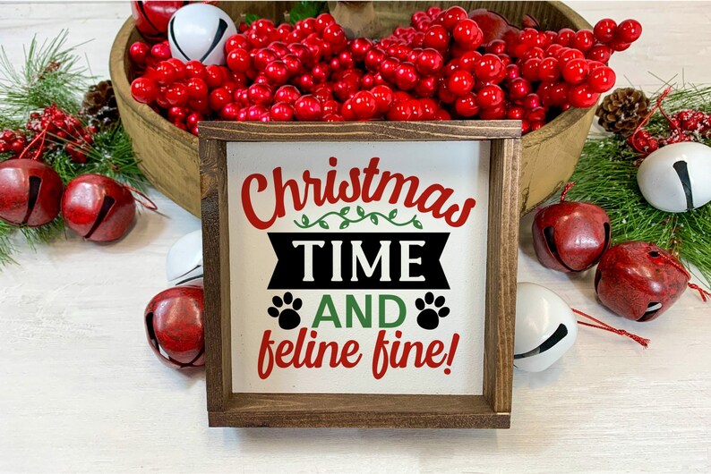 May include: A wooden sign with the text "Christmas Time And Feline Fine!" in red, green, and black lettering. The sign is surrounded by red and white jingle bells.