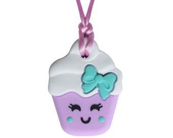 May include: A purple cupcake-shaped silicone necklace with a white frosting top, a turquoise bow, and a smiling face. The necklace is on a pink cord.