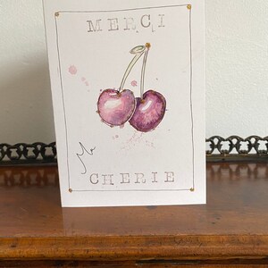 May include: A white card with gold accents features a watercolor illustration of two purple cherries with green stems. The card says "Merci, ma Cherie" in black text.