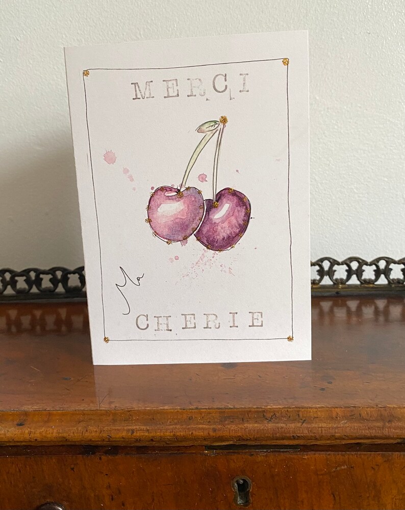 May include: A white card with gold accents features a watercolor illustration of two purple cherries with green stems. The card says "Merci, ma Cherie" in black text.