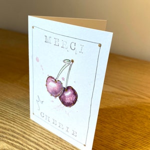 May include: A white card with gold glitter accents. The card features a watercolor illustration of two purple cherries with green stems and gold glitter accents. The text "MERCI, MA CHERIE" is printed in black on the card.