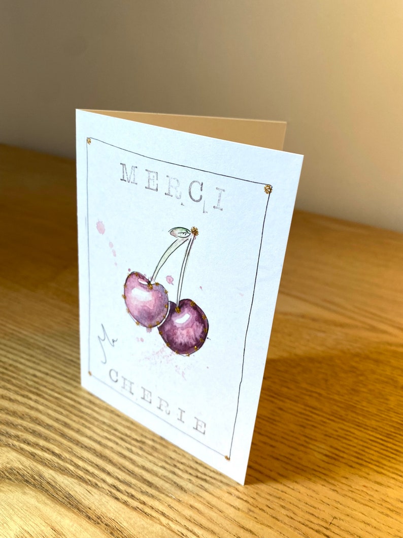 May include: A white card with gold glitter accents. The card features a watercolor illustration of two purple cherries with green stems and gold glitter accents. The text "MERCI, MA CHERIE" is printed in black on the card.