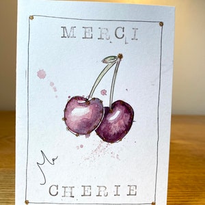 May include: A white card with a gold border and the words "MERCI MA CHERIE" in black lettering. A watercolor painting of two purple cherries with green stems is in the center of the card.