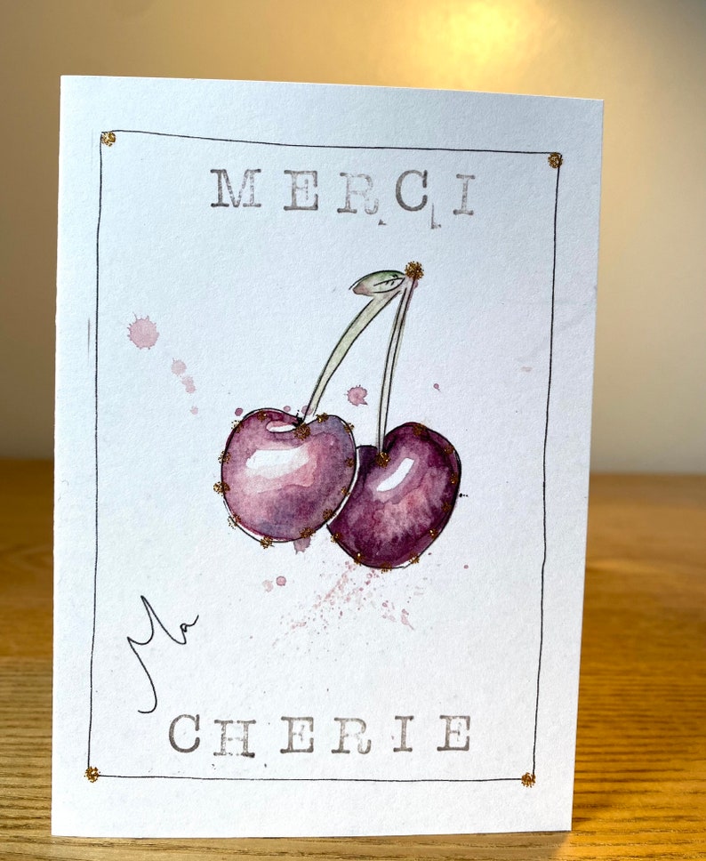 May include: A white card with a gold border and the words "MERCI MA CHERIE" in black lettering. A watercolor painting of two purple cherries with green stems is in the center of the card.