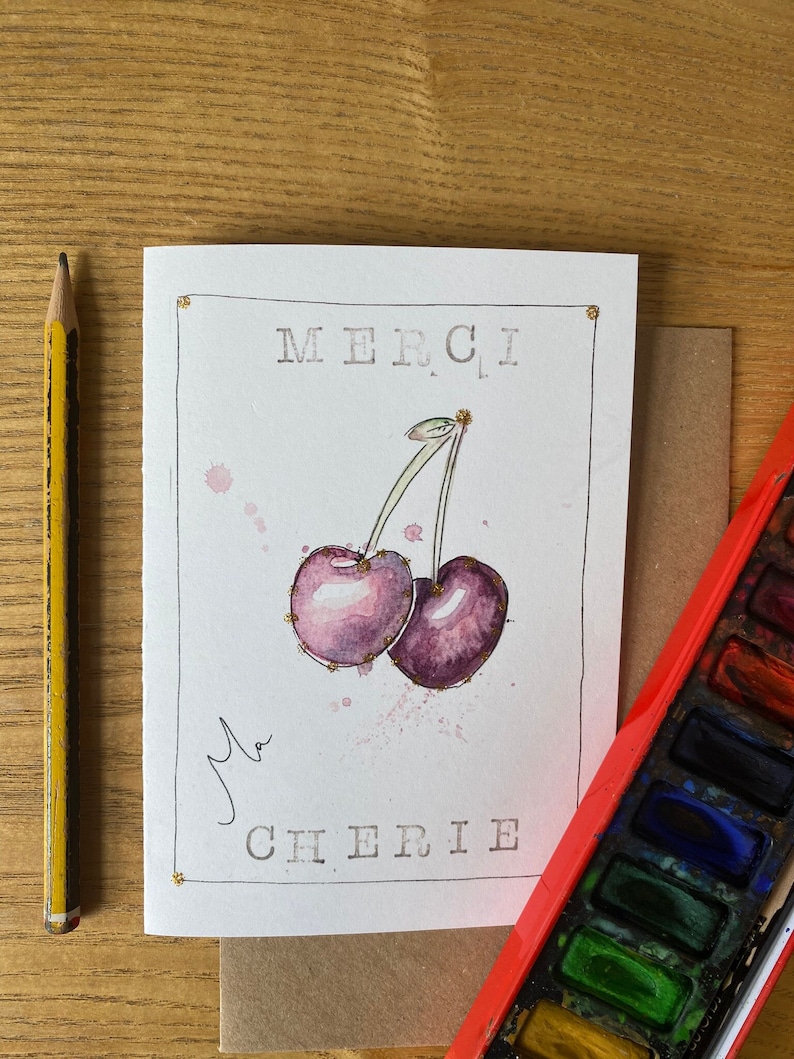 May include: A white card with gold accents features a watercolor illustration of two purple cherries with gold glitter accents. The card says "MERCI, Ma CHERIE" in black text.